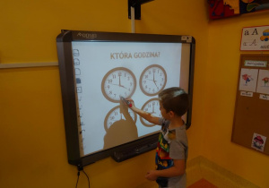 Chłopiec stoi przy tablicy interaktywnej na której są cztery tarcze zegarów, wskaźnikiem wskazuje na jeden z nich i odczytuje godzinę 4.